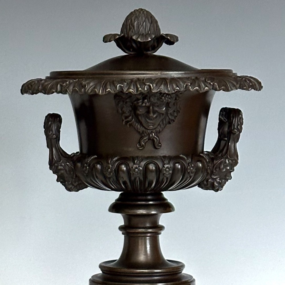 A pair of patinated dark bronze covered urns featuring classical mask motifs, acanthus leaf moulding, twin handles, and pinecone finials on round stepped bases. View V