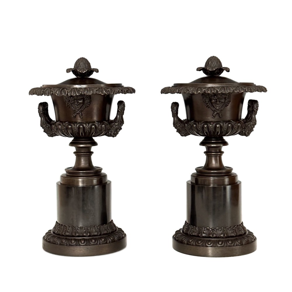 A pair of patinated dark bronze covered urns featuring classical mask motifs, acanthus leaf moulding, twin handles, and pinecone finials on round stepped bases. View VI