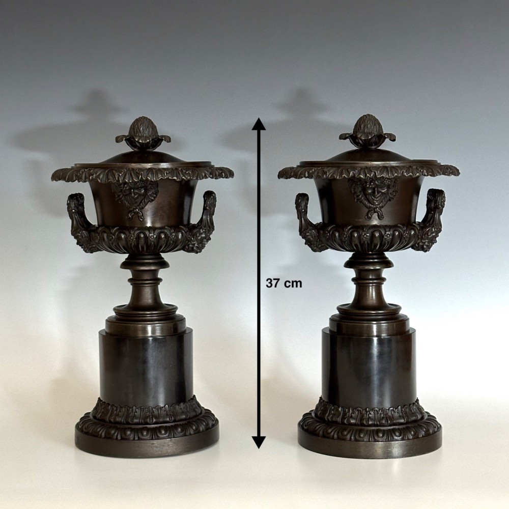A pair of patinated dark bronze covered urns featuring classical mask motifs, acanthus leaf moulding, twin handles, and pinecone finials on round stepped bases. View VII