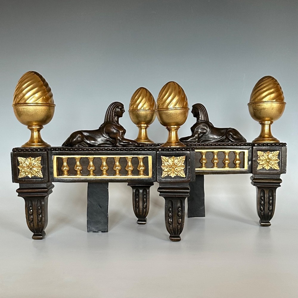 A pair of Regency bronze and ormolu chenets featuring sculpted bronze sphinxes, gilded balustrades, pinecone finials, and fluted legs.