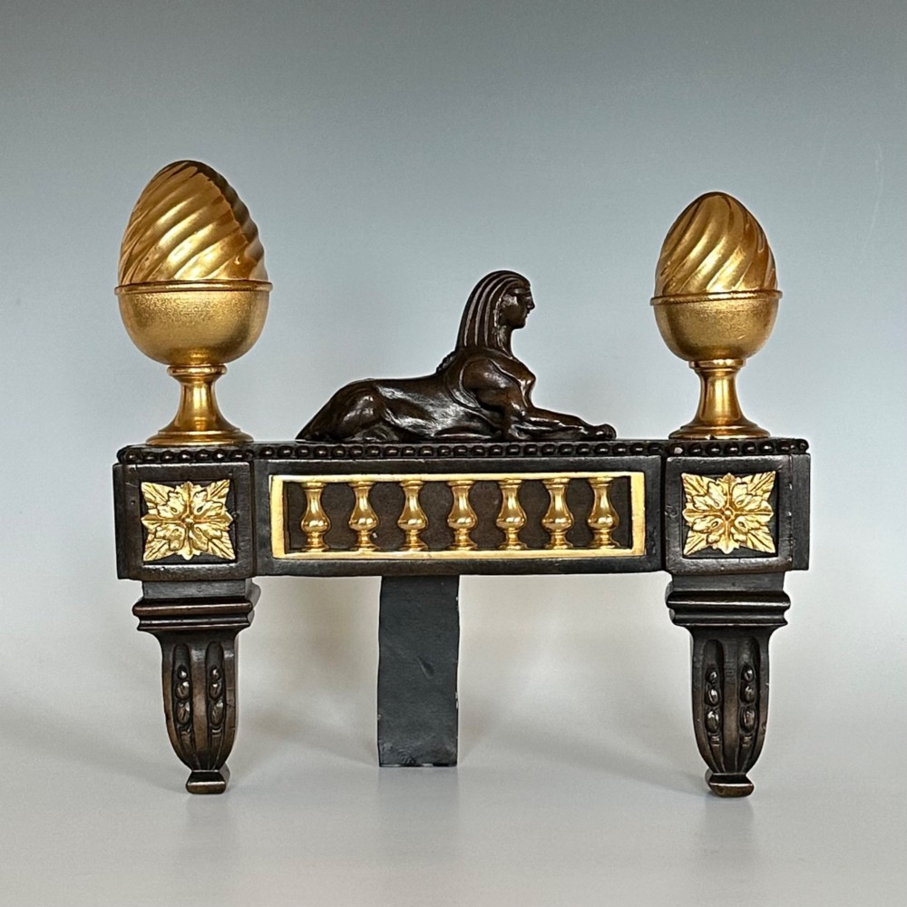 A pair of Regency bronze and ormolu chenets featuring sculpted bronze sphinxes, gilded balustrades, pinecone finials, and fluted legs. View II