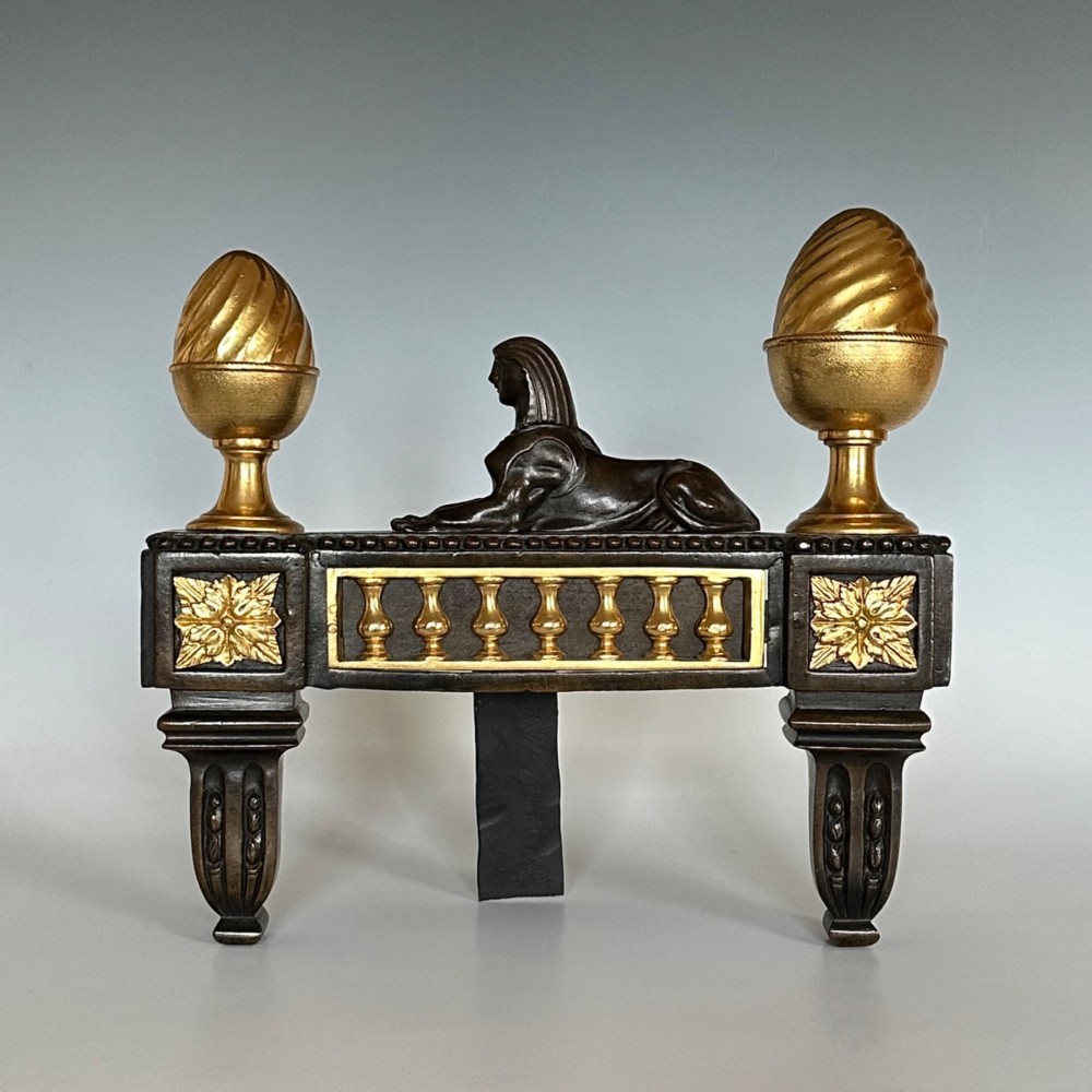 A pair of Regency bronze and ormolu chenets featuring sculpted bronze sphinxes, gilded balustrades, pinecone finials, and fluted legs. View III