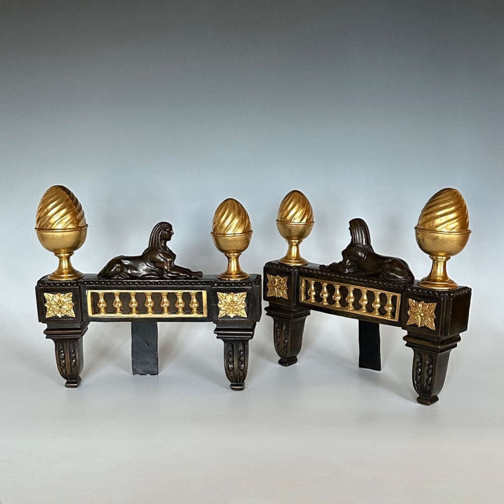 A pair of Regency bronze and ormolu chenets featuring sculpted bronze sphinxes, gilded balustrades, pinecone finials, and fluted legs. View V