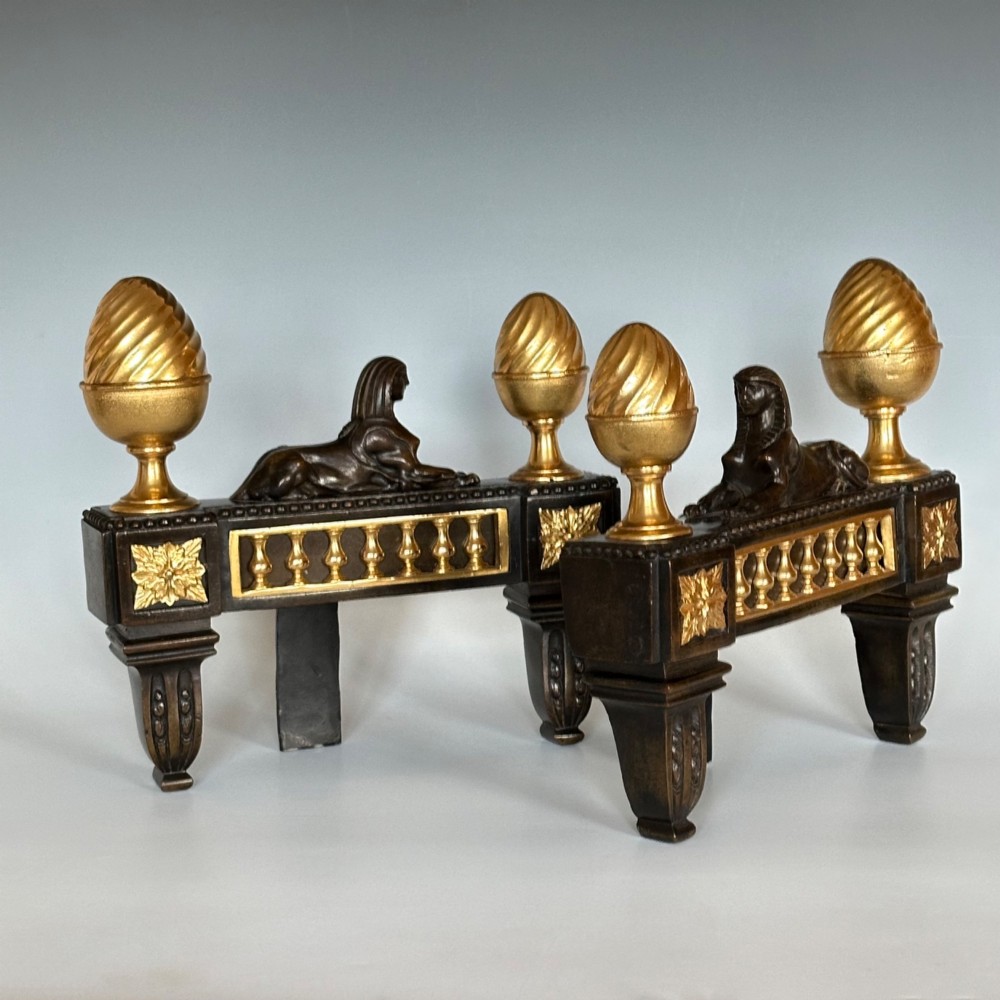 A pair of Regency bronze and ormolu chenets featuring sculpted bronze sphinxes, gilded balustrades, pinecone finials, and fluted legs. View VI