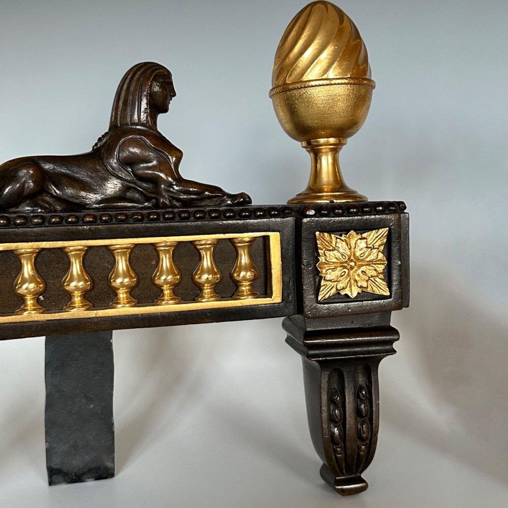 A pair of Regency bronze and ormolu chenets featuring sculpted bronze sphinxes, gilded balustrades, pinecone finials, and fluted legs. View VIII