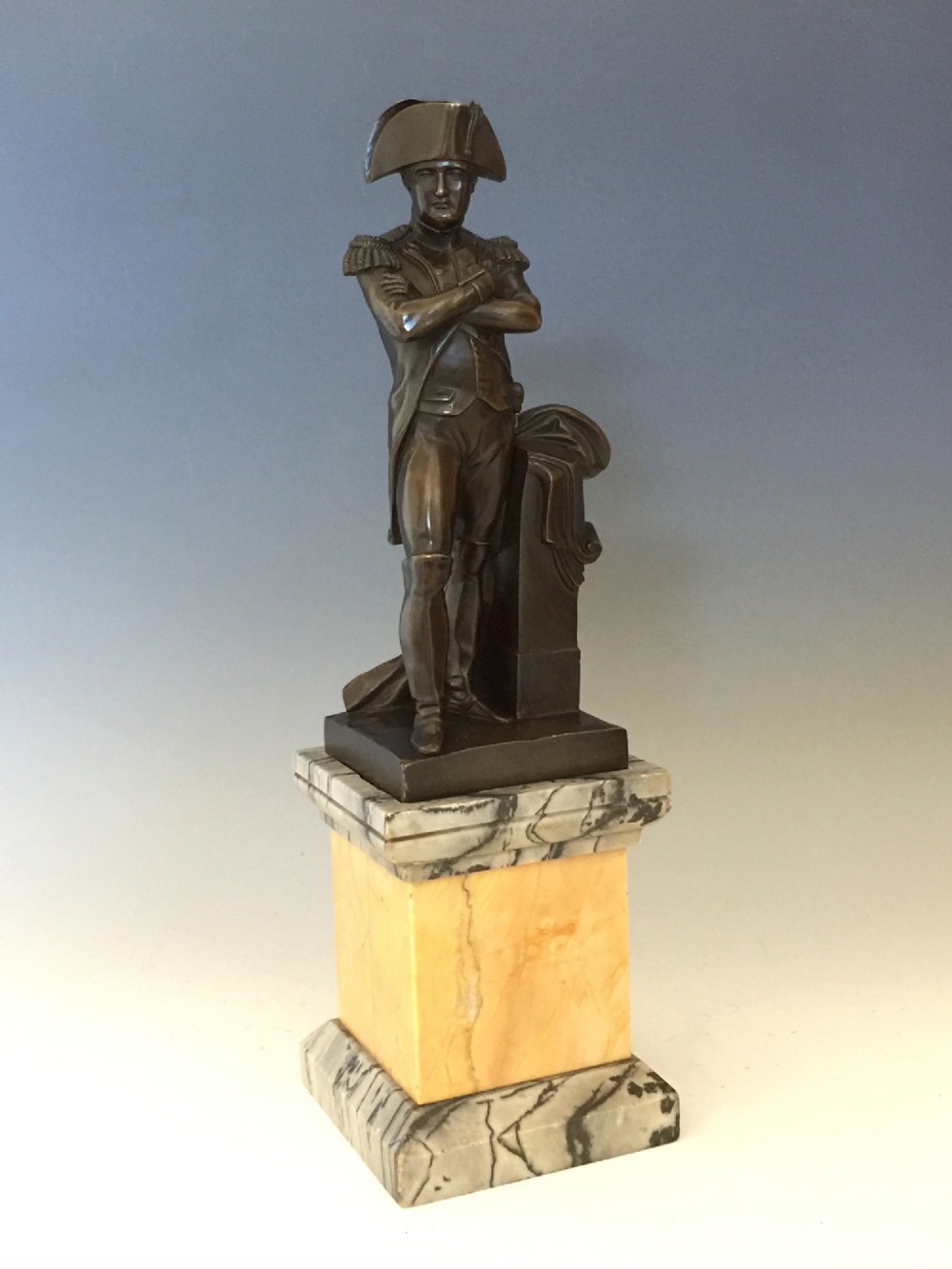 A nineteenth-century bronze figure of Napoleon Bonaparte in full military uniform standing with arms crossed on a yellow and grey marble plinth.