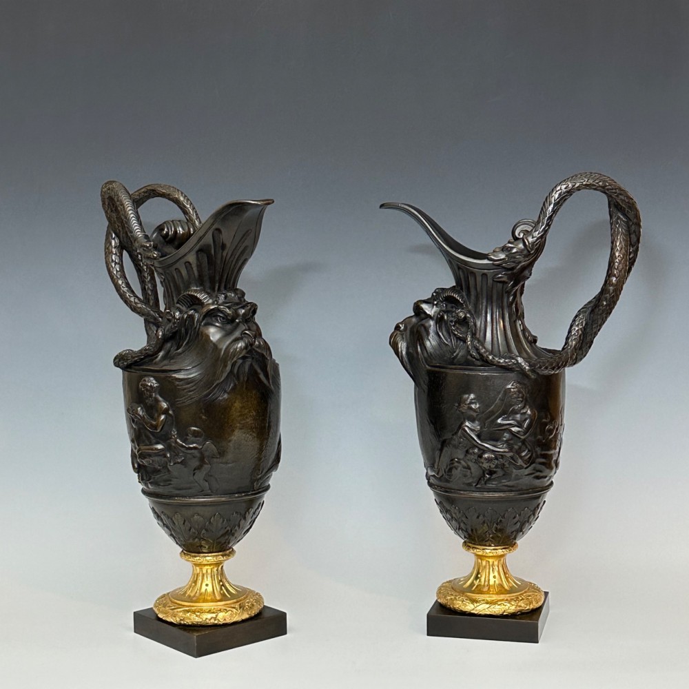 A pair of dark patinated bronze French ewers featuring relief carvings of classical mythological figures, serpent handles, satyr masks, and gilt bronze bases.