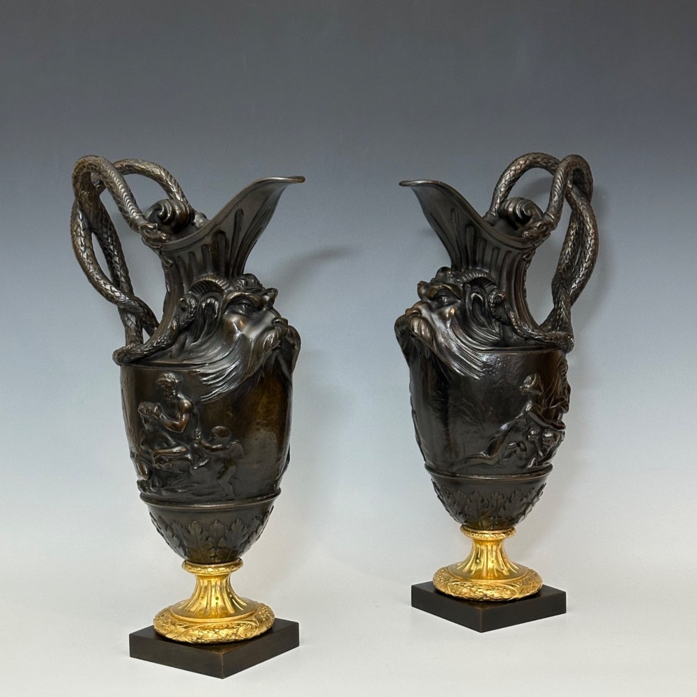 A pair of dark patinated bronze French ewers featuring relief carvings of classical mythological figures, serpent handles, satyr masks, and gilt bronze bases. View X