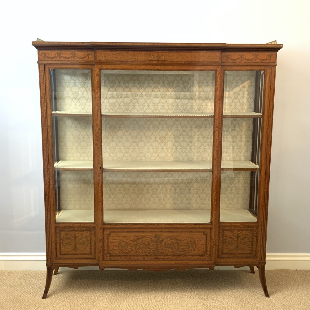 A late Victorian satinwood display cabinet featuring three glazed doors, detailed marquetry inlay, fabric-lined interior shelves, and splayed legs.