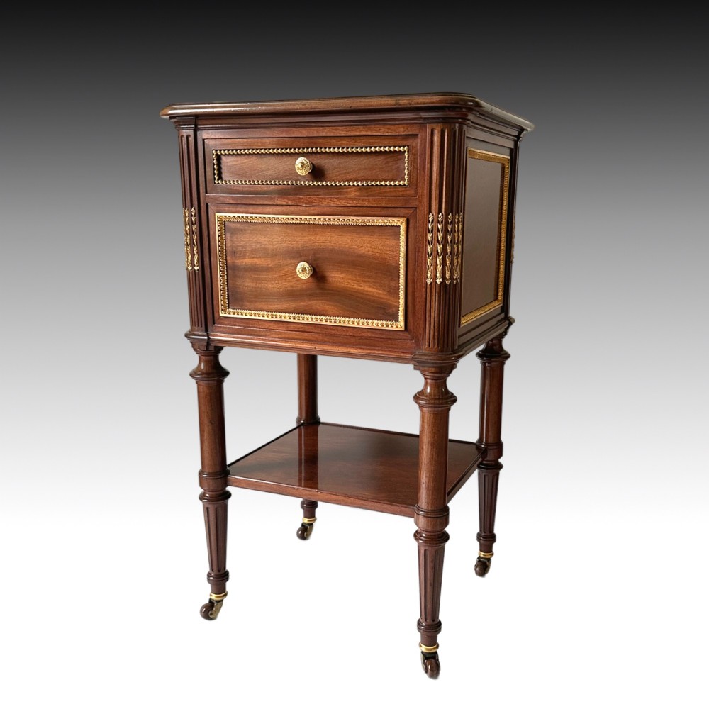A French Louis XVI style mahogany bedside cabinet featuring two drawers framed with beaded ormolu trim, fluted corner columns, a lower tier shelf, and turned legs with brass castors.