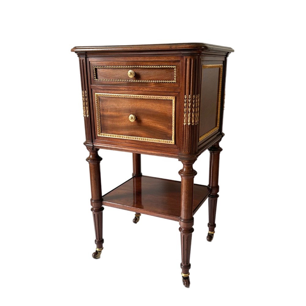 A French Louis XVI style mahogany bedside cabinet featuring two drawers framed with beaded ormolu trim, fluted corner columns, a lower tier shelf, and turned legs with brass castors. View II