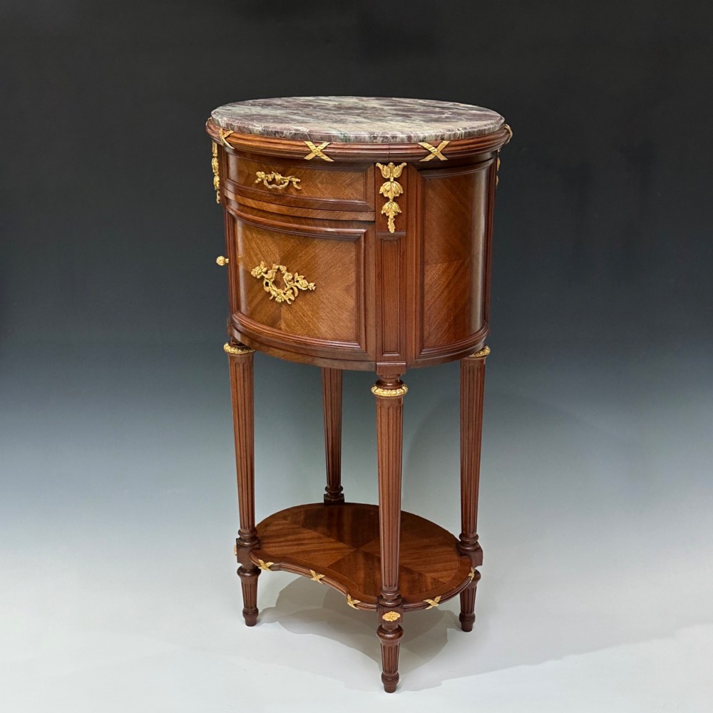 An oval French mahogany table de nuit featuring a grey marble top, gilt-bronze mounts, single drawer, cupboard door, and fluted legs with an undertier.