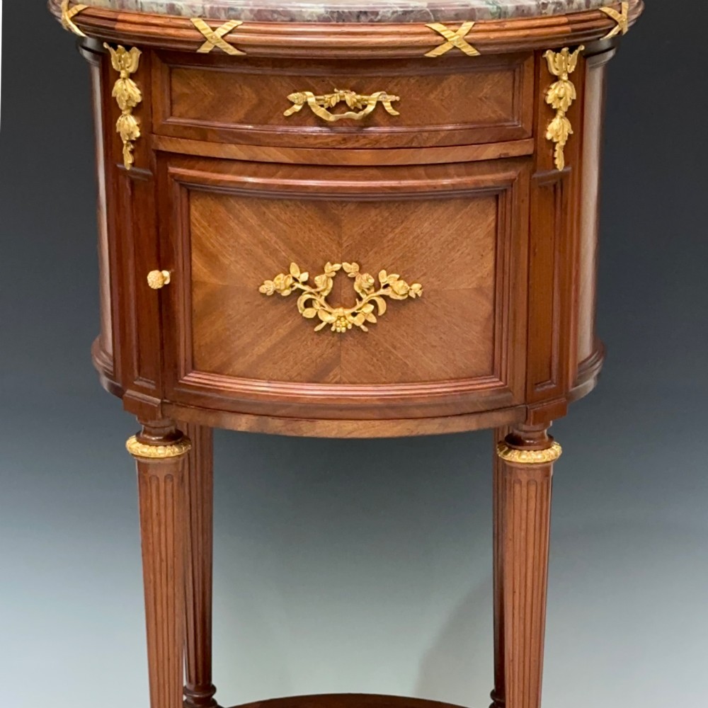 An oval French mahogany table de nuit featuring a grey marble top, gilt-bronze mounts, single drawer, cupboard door, and fluted legs with an undertier. View II