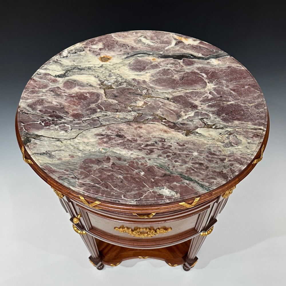 An oval French mahogany table de nuit featuring a grey marble top, gilt-bronze mounts, single drawer, cupboard door, and fluted legs with an undertier. View IV