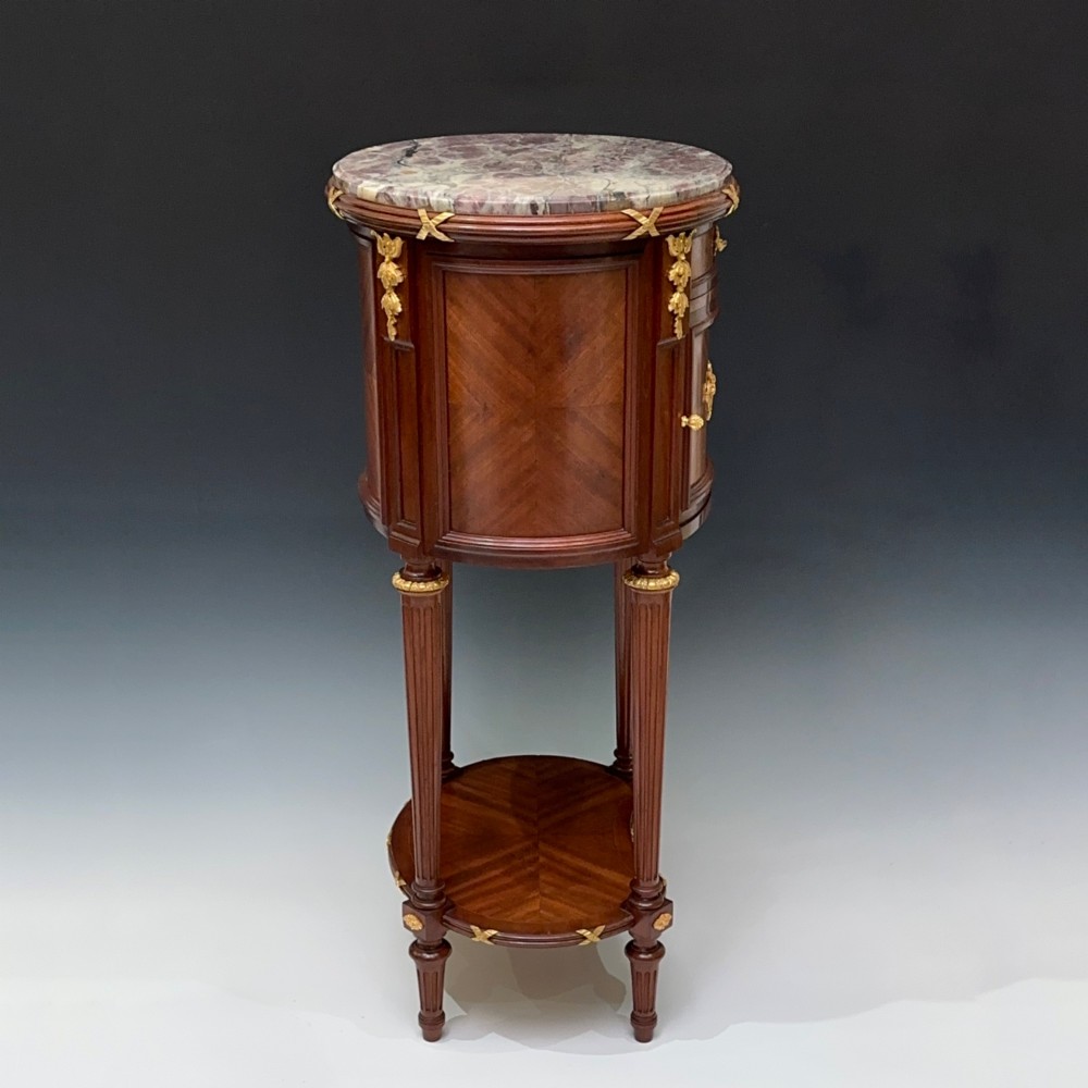 An oval French mahogany table de nuit featuring a grey marble top, gilt-bronze mounts, single drawer, cupboard door, and fluted legs with an undertier. View V