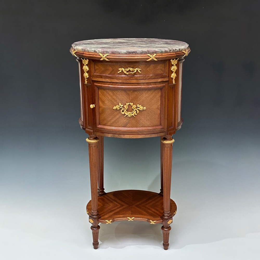 An oval French mahogany table de nuit featuring a grey marble top, gilt-bronze mounts, single drawer, cupboard door, and fluted legs with an undertier. View VI