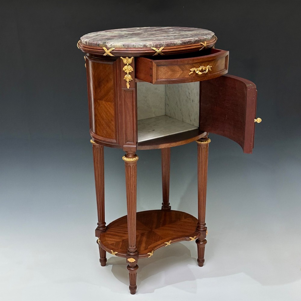 An oval French mahogany table de nuit featuring a grey marble top, gilt-bronze mounts, single drawer, cupboard door, and fluted legs with an undertier. View VII