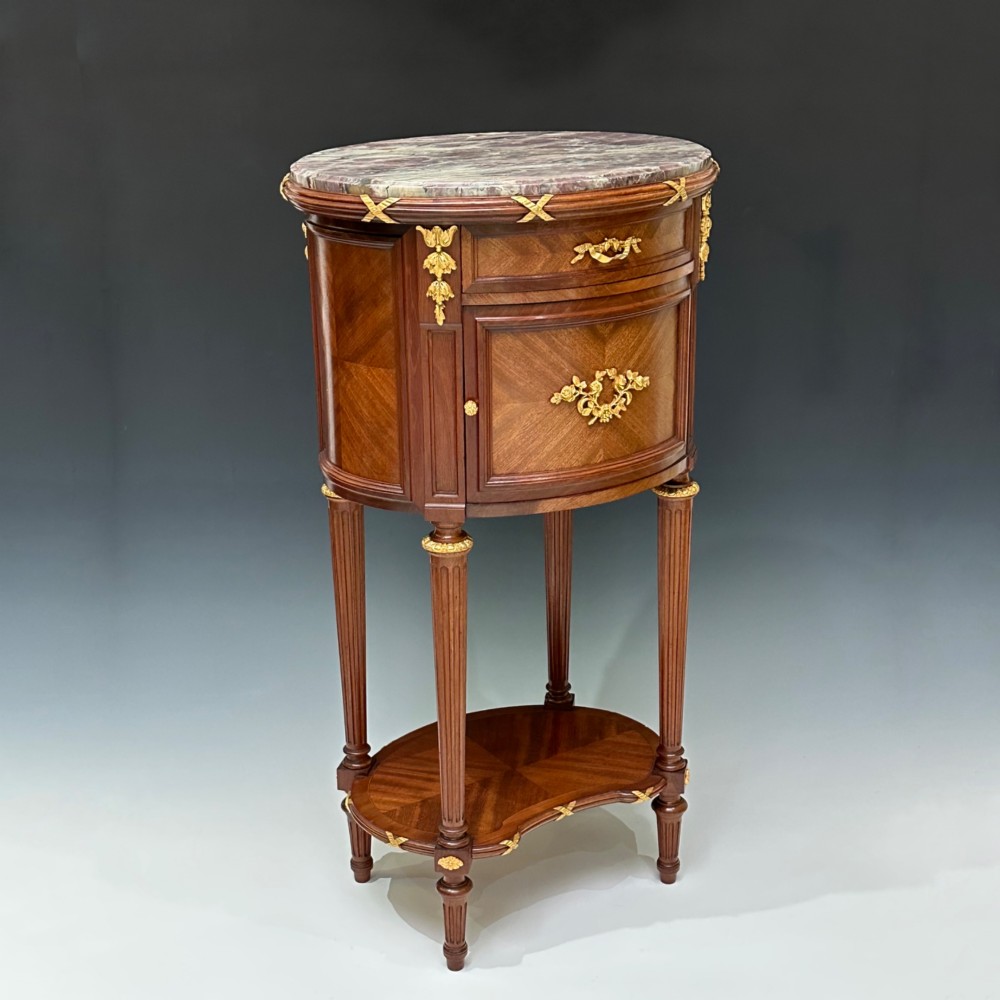 An oval French mahogany table de nuit featuring a grey marble top, gilt-bronze mounts, single drawer, cupboard door, and fluted legs with an undertier. View X