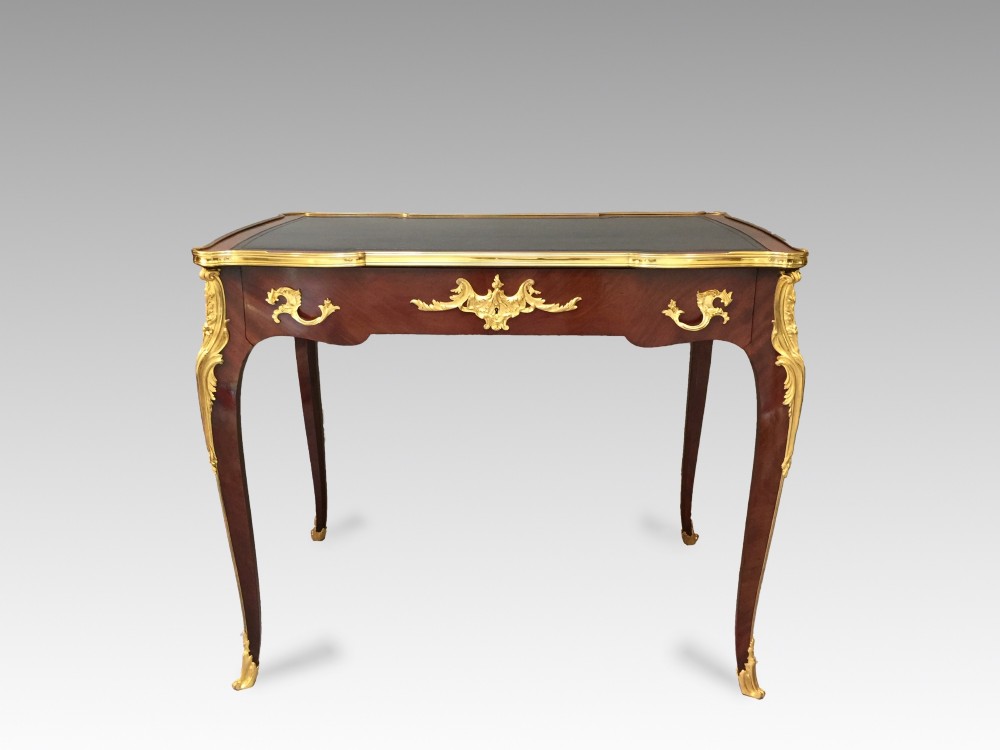A French Louis XV style bureau plat crafted from dark polished wood with serpentine cabriole legs, elaborate gilt-bronze ormolu mounts, and a black leather writing top. View IV