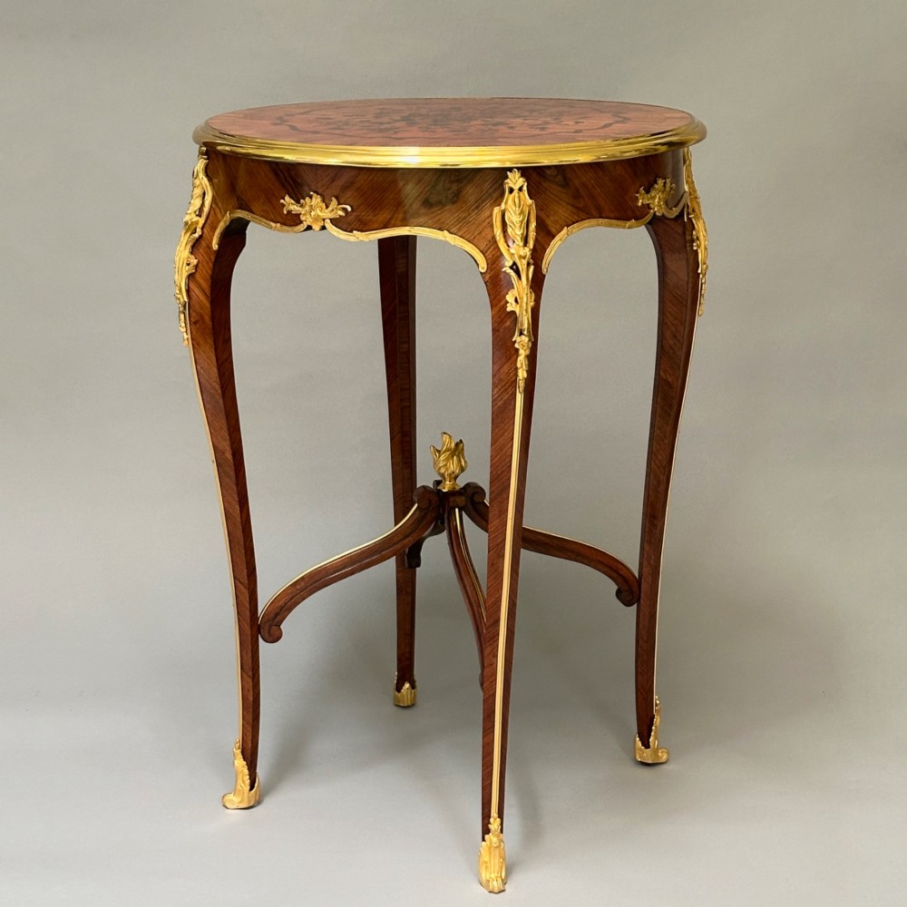 A round French kingwood occasional table featuring floral marquetry inlay, gilded ormolu mounts, slender cabriole legs, and a central stretcher with a carved finial.