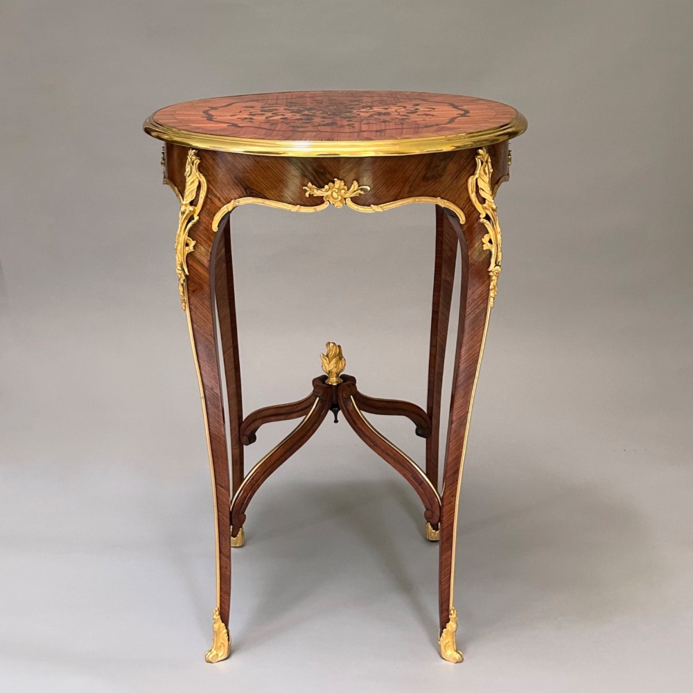 A round French kingwood occasional table featuring floral marquetry inlay, gilded ormolu mounts, slender cabriole legs, and a central stretcher with a carved finial. View V