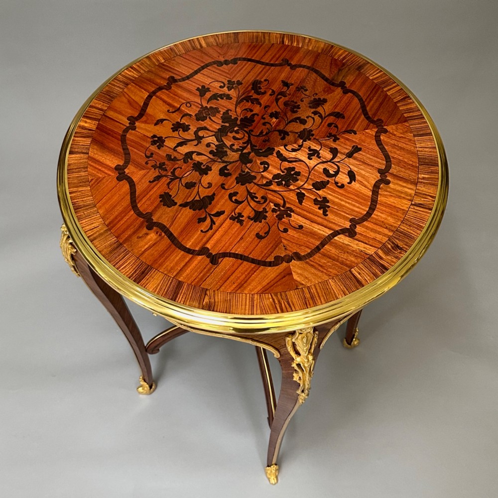 A round French kingwood occasional table featuring floral marquetry inlay, gilded ormolu mounts, slender cabriole legs, and a central stretcher with a carved finial. View VI
