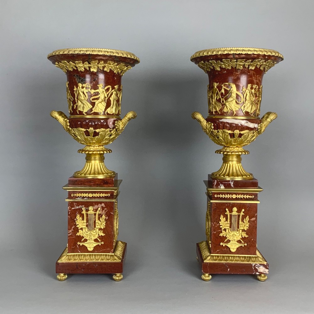 A pair of French Empire style red marble urns featuring gilded bronze classical figure reliefs and mounted on square marble pedestals with lyre details. View VI