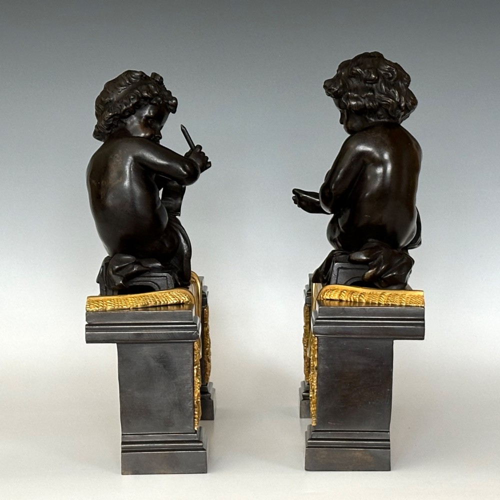 A pair of dark patinated bronze putti figures seated atop rectangular bases featuring gilt drapery and ornate floral side appliques. View V