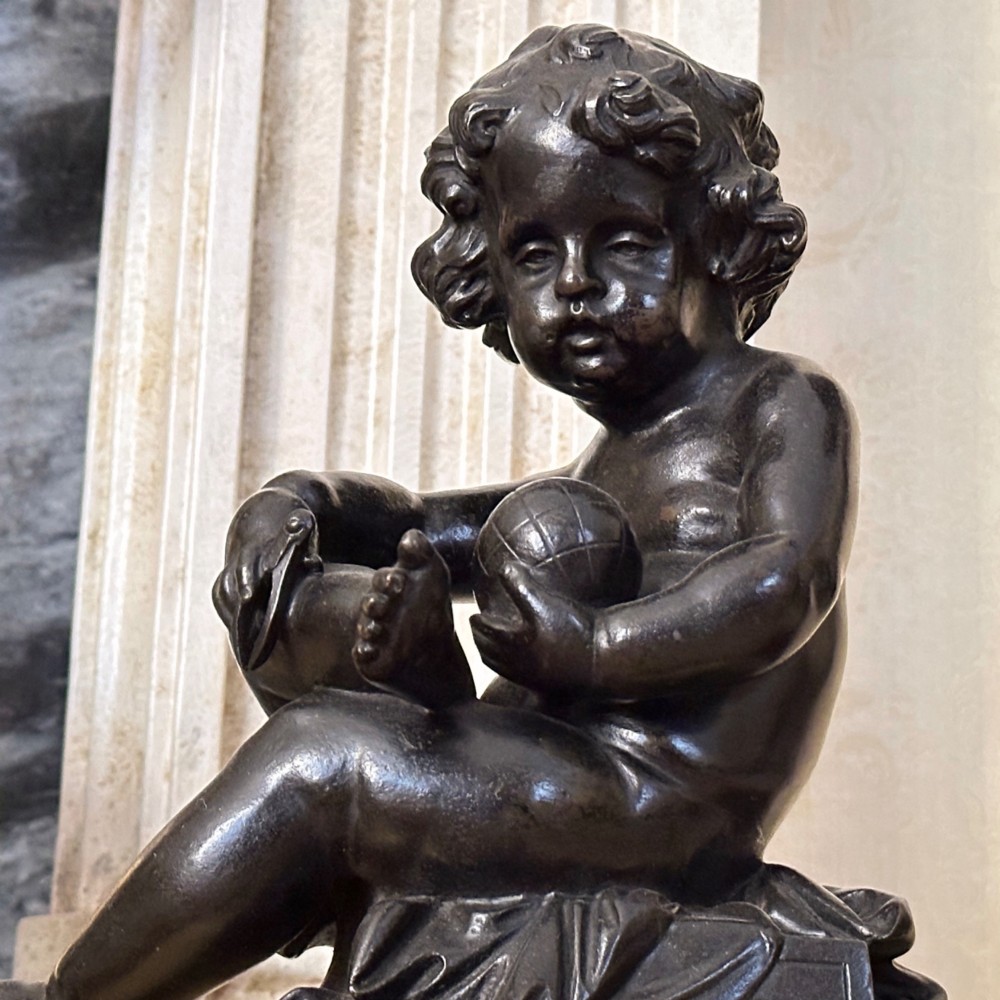 A pair of dark patinated bronze putti figures seated atop rectangular bases featuring gilt drapery and ornate floral side appliques. View VI
