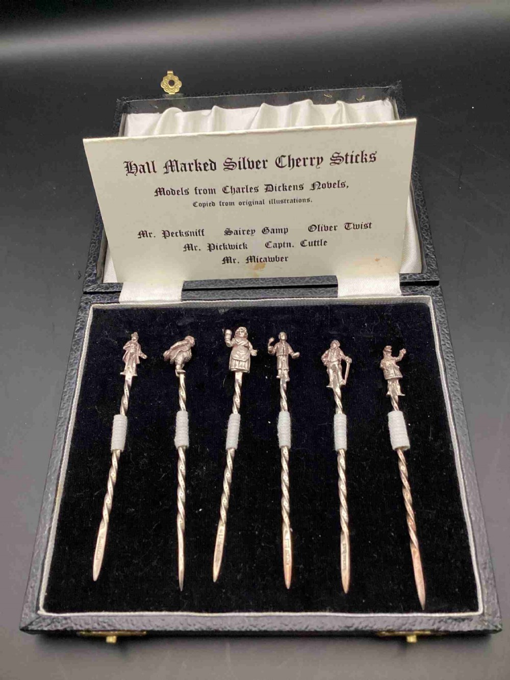 A set of six sterling silver cherry sticks with detailed Charles Dickens character finials resting in a black fitted velvet case.