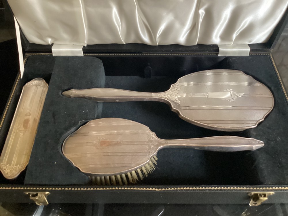 An antique hallmarked silver dressing table set including a hand mirror and two brushes with engine-turned detailing, resting inside a black velvet-lined presentation box.