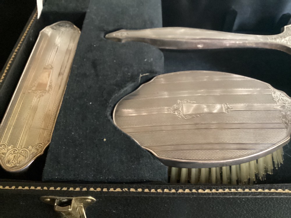 An antique hallmarked silver dressing table set including a hand mirror and two brushes with engine-turned detailing, resting inside a black velvet-lined presentation box. View III