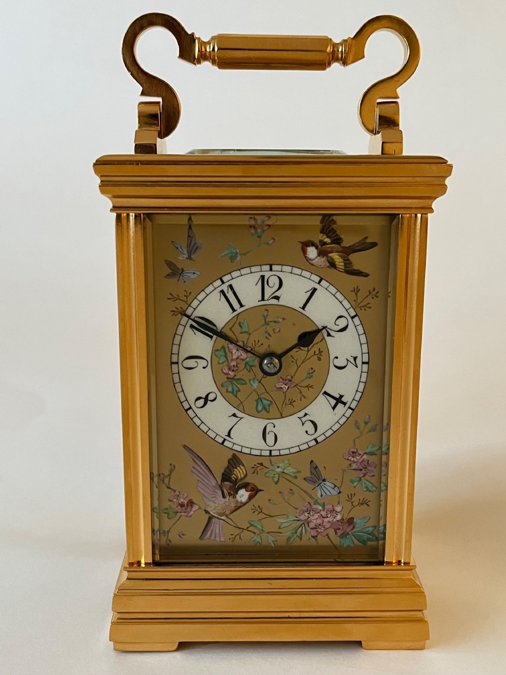 An antique French gilt brass carriage clock featuring a top folding handle, glass side panels, and a hand-painted dial with bird and floral motifs.