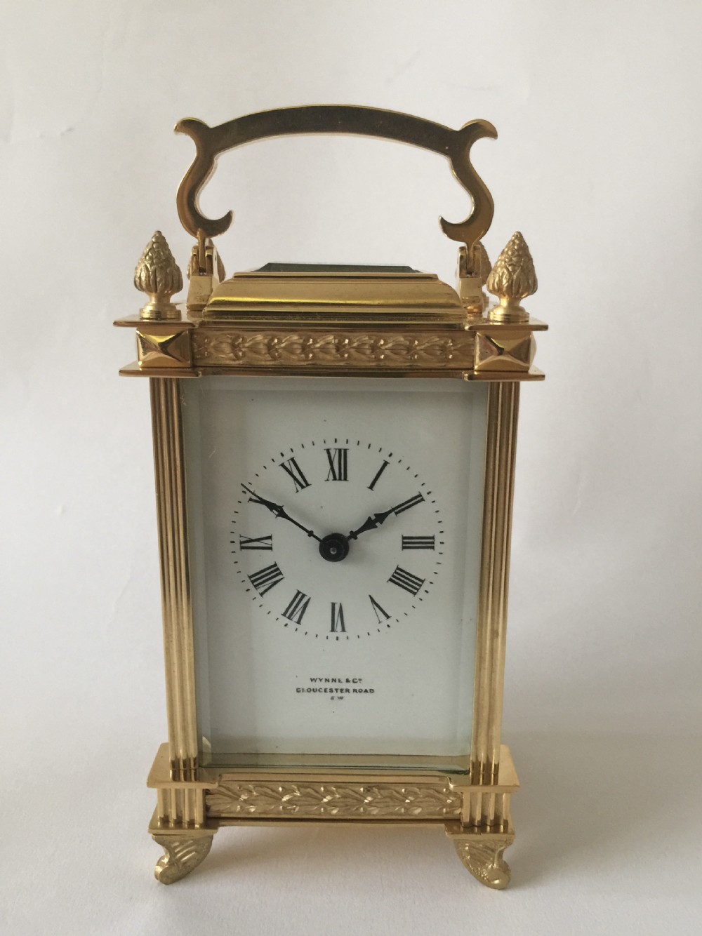 A rectangular gilt brass carriage clock featuring an enamel dial with Roman numerals, fluted corner columns, foliage frieze detailing, and a top carrying handle.