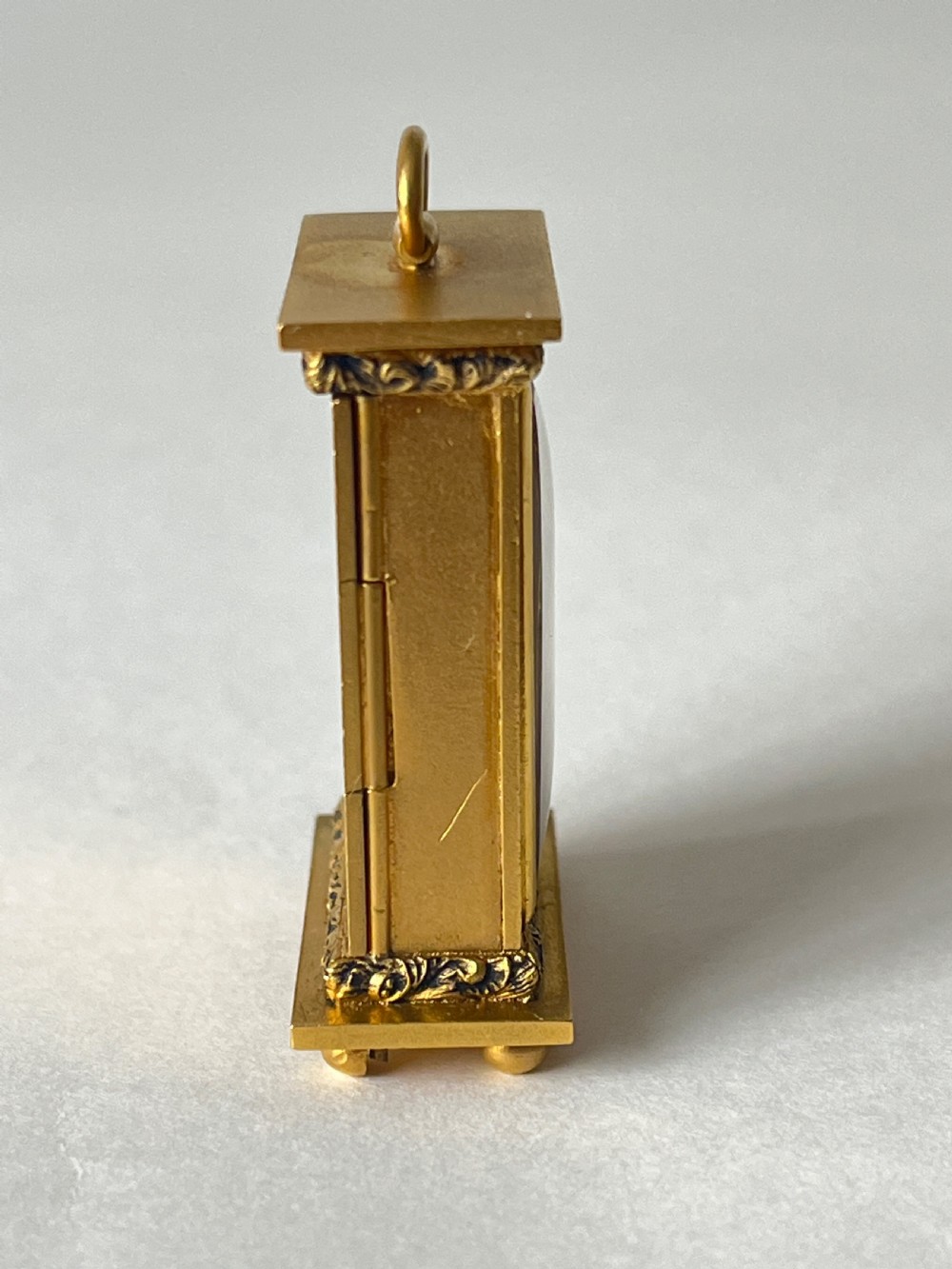 A small rectangular gilt brass Art Deco carriage clock featuring a top loop handle, chased floral borders, ball feet, and a golden guilloche dial with dark stylized numbers. View III
