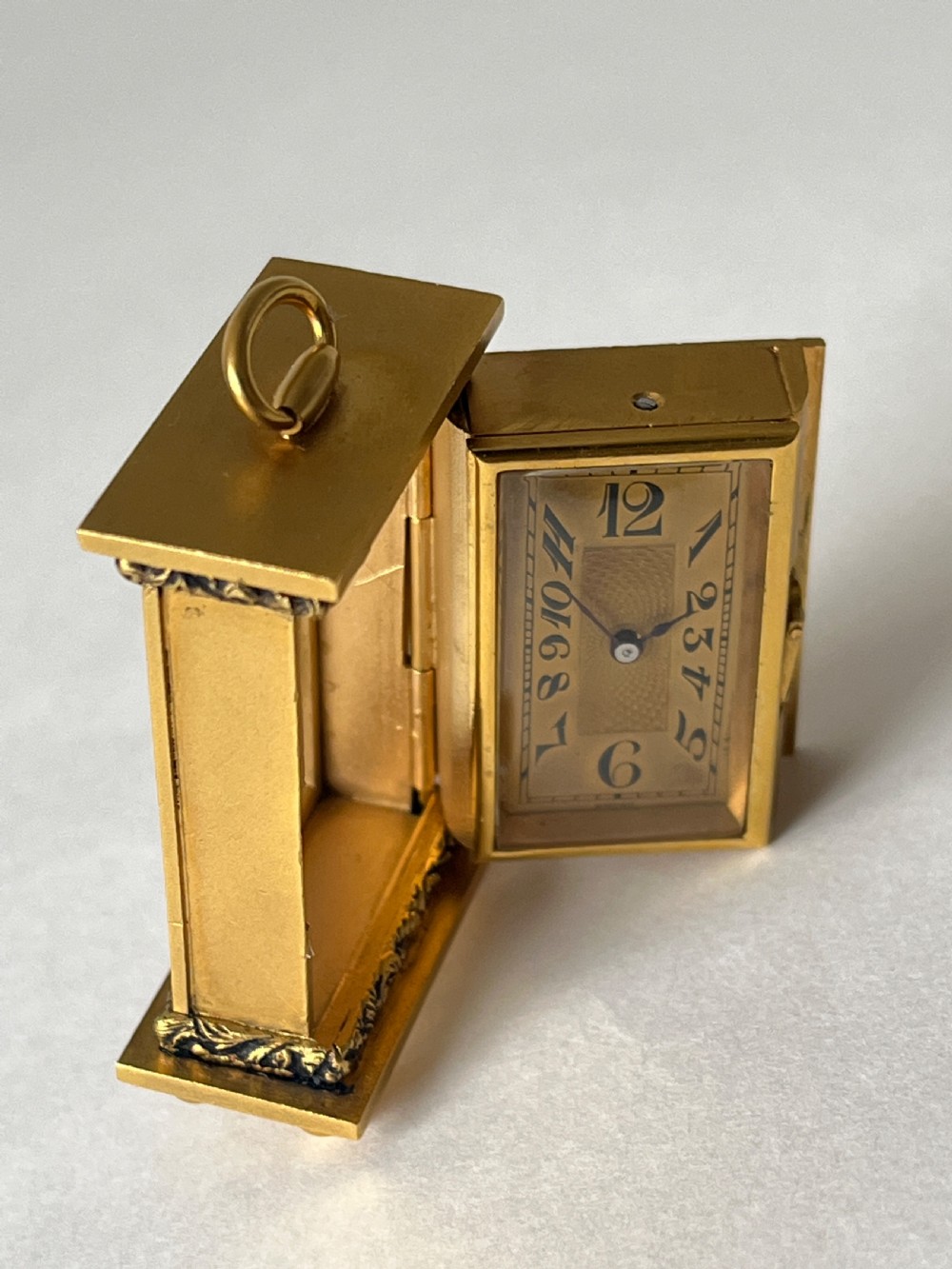A small rectangular gilt brass Art Deco carriage clock featuring a top loop handle, chased floral borders, ball feet, and a golden guilloche dial with dark stylized numbers. View IV