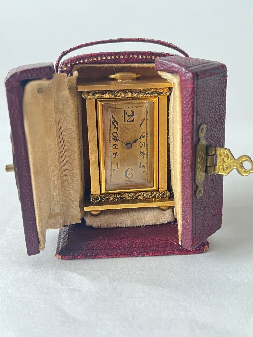 A small rectangular gilt brass Art Deco carriage clock featuring a top loop handle, chased floral borders, ball feet, and a golden guilloche dial with dark stylized numbers. View VII