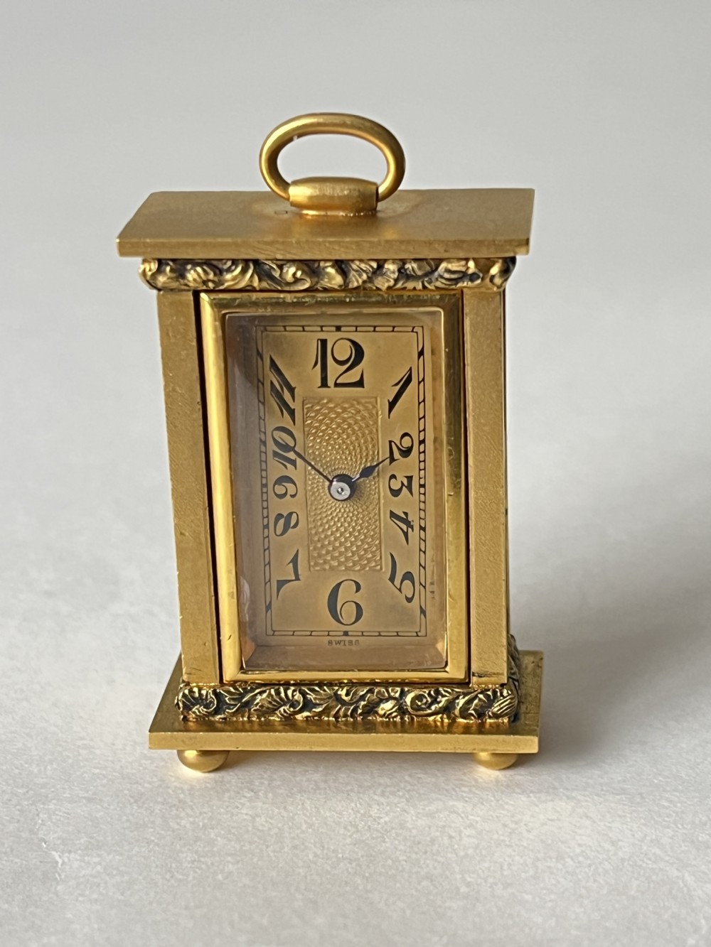 A small rectangular gilt brass Art Deco carriage clock featuring a top loop handle, chased floral borders, ball feet, and a golden guilloche dial with dark stylized numbers.