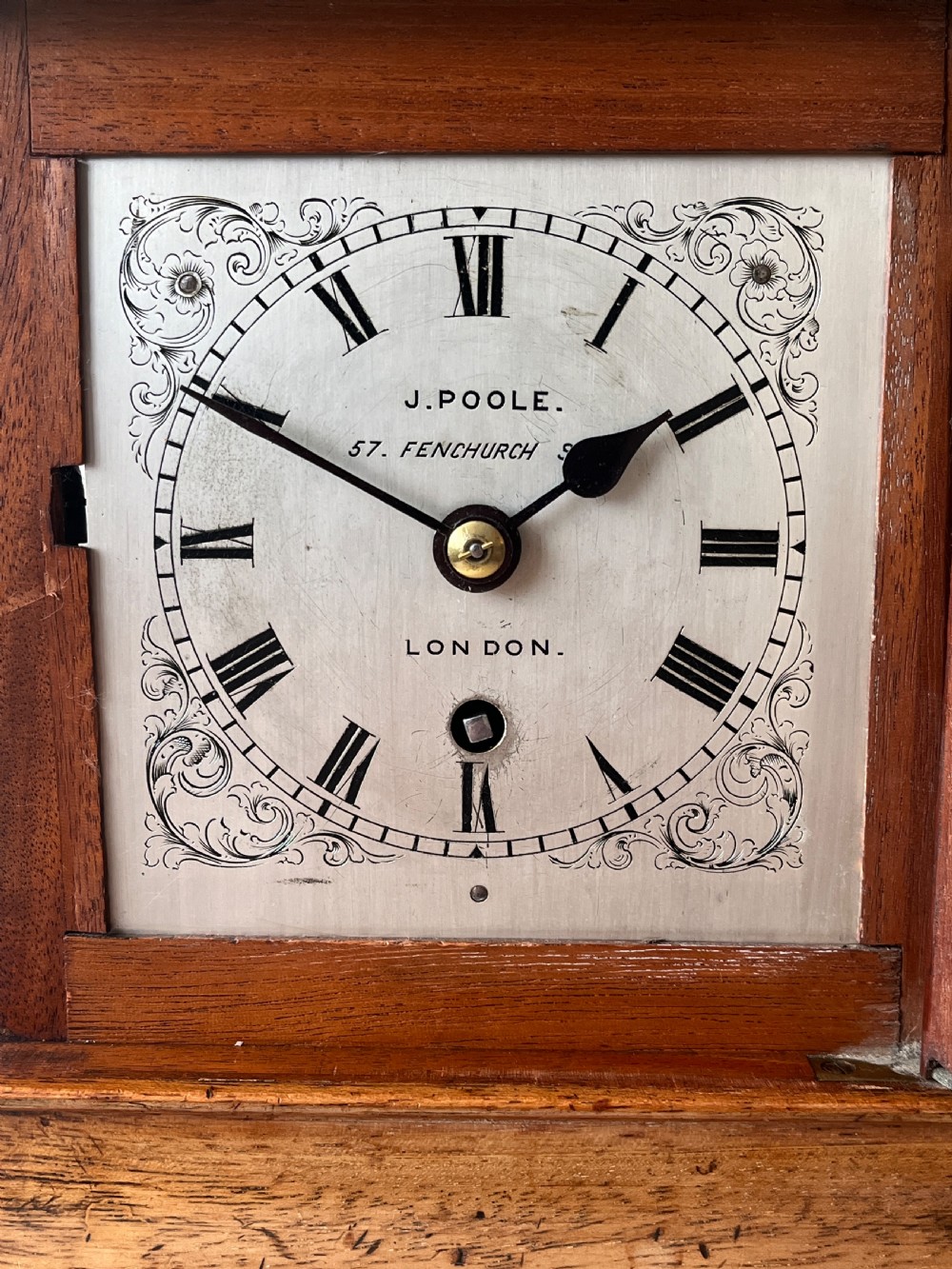 A Victorian mahogany library clock featuring a square timber case with a silvered engraved dial, black Roman numerals, and a key visible on the side. View VI