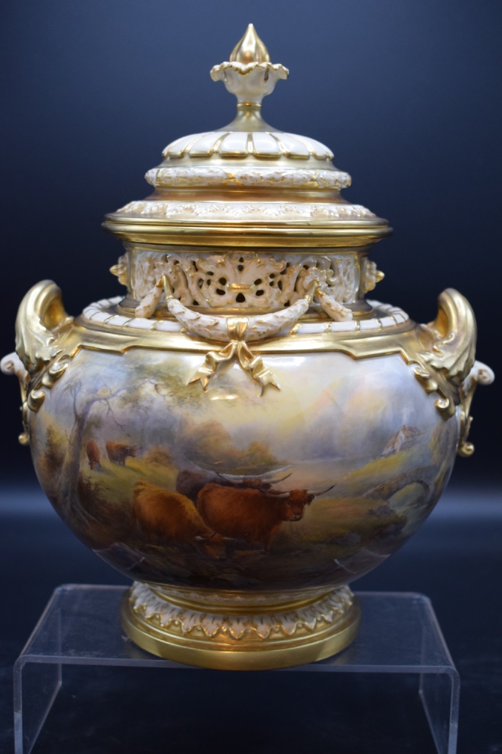 A gilded porcelain pot pourri vase with a pierced cover, twin scroll handles, and a painted landscape featuring Highland cattle.