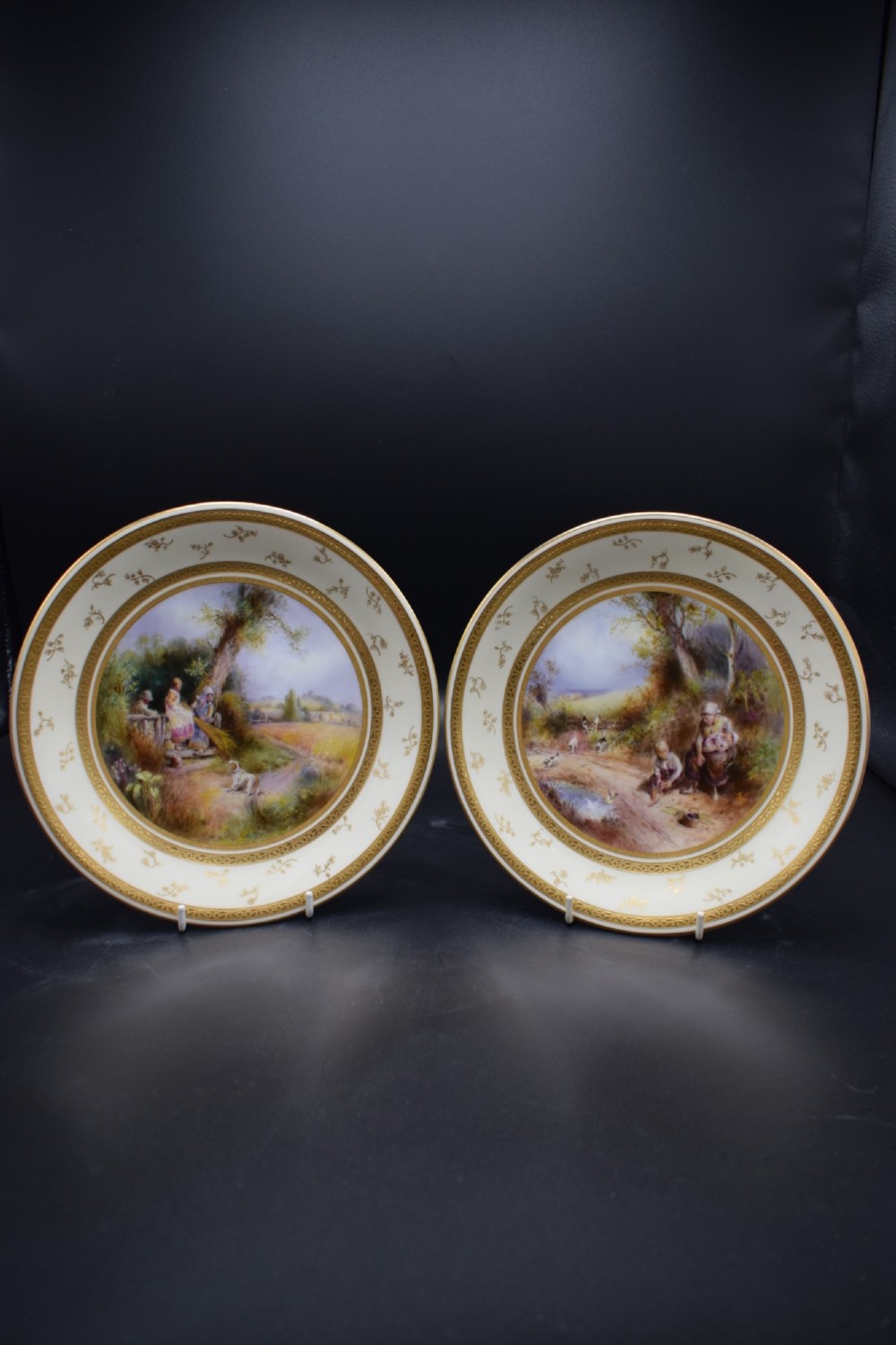 A pair of circular porcelain plates decorated with hand-painted pastoral landscapes and framed by wide cream borders with intricate gilded detailing.