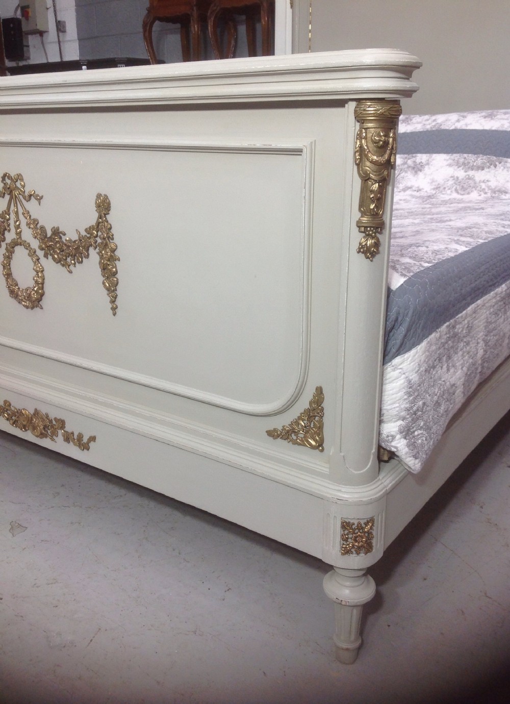 An off-white painted French Neoclassical bed frame featuring a tall rectangular headboard, fluted turned legs, and intricate brass swag mounts on the footboard. View II