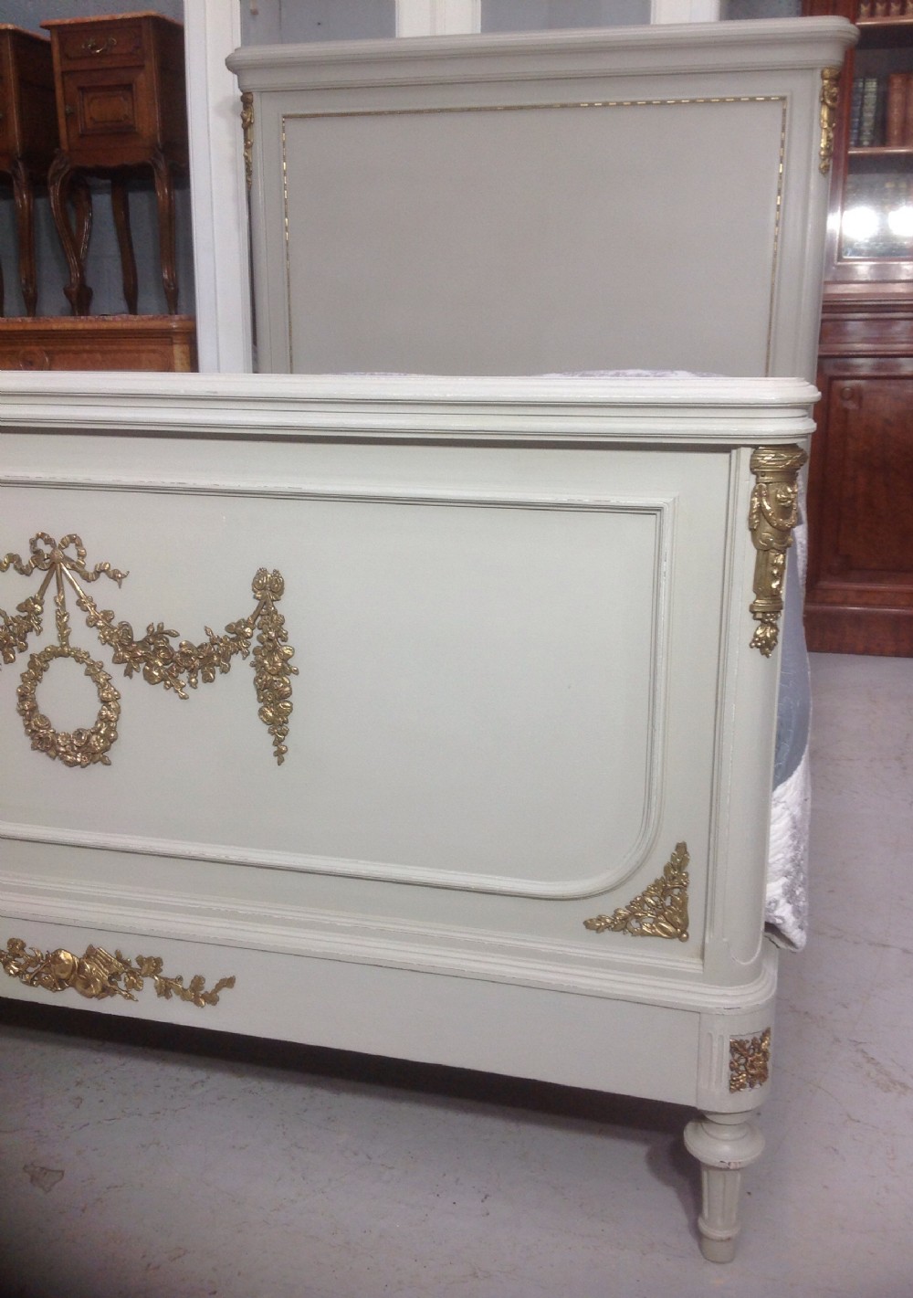 An off-white painted French Neoclassical bed frame featuring a tall rectangular headboard, fluted turned legs, and intricate brass swag mounts on the footboard. View III