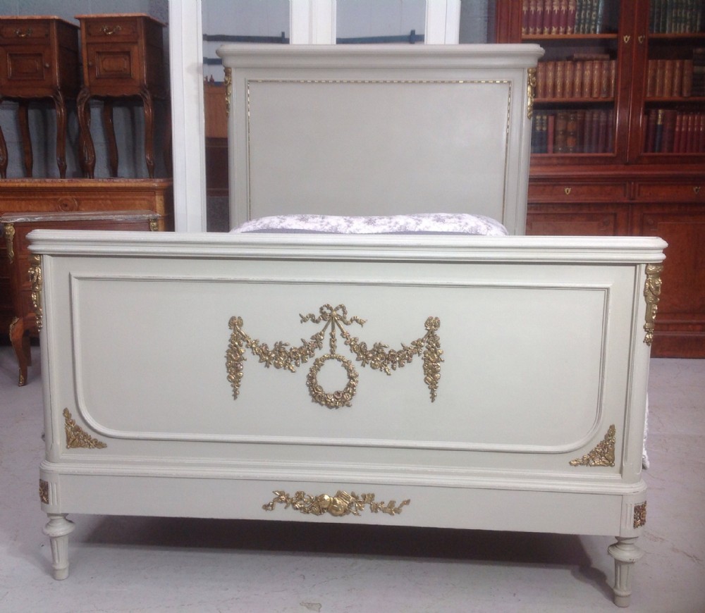 An off-white painted French Neoclassical bed frame featuring a tall rectangular headboard, fluted turned legs, and intricate brass swag mounts on the footboard. View IV