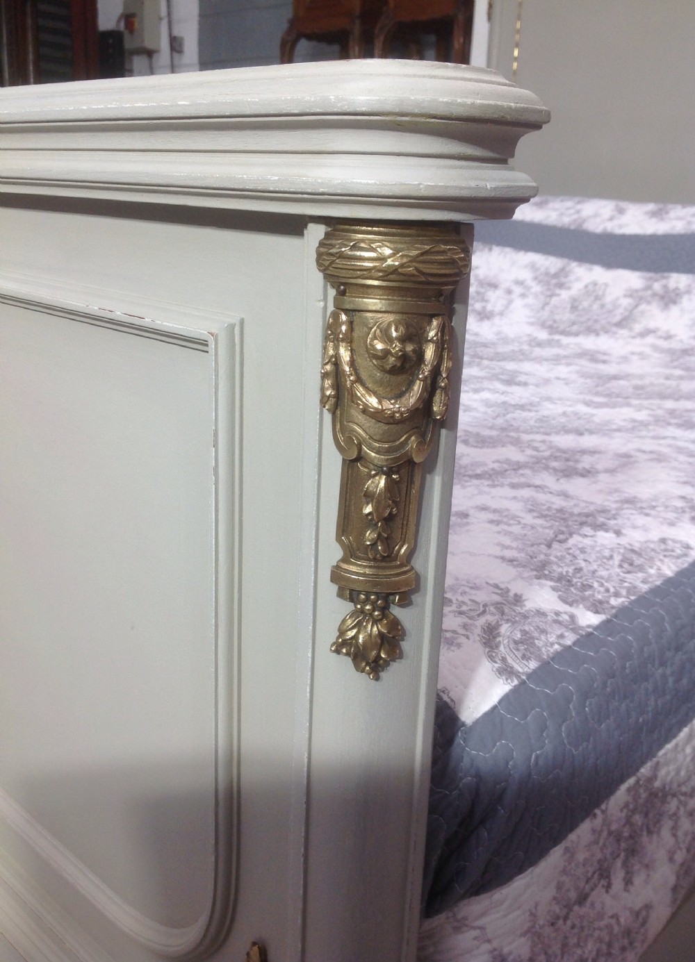 An off-white painted French Neoclassical bed frame featuring a tall rectangular headboard, fluted turned legs, and intricate brass swag mounts on the footboard. View VIII
