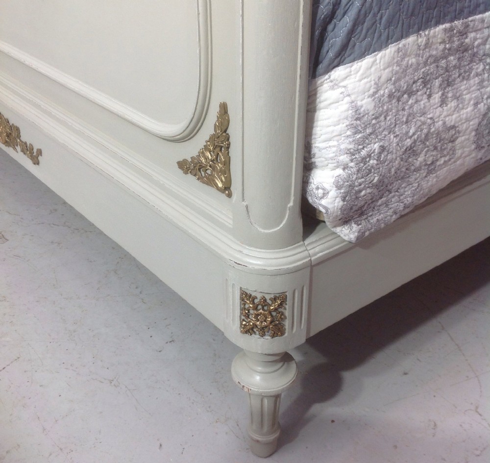 An off-white painted French Neoclassical bed frame featuring a tall rectangular headboard, fluted turned legs, and intricate brass swag mounts on the footboard. View IX