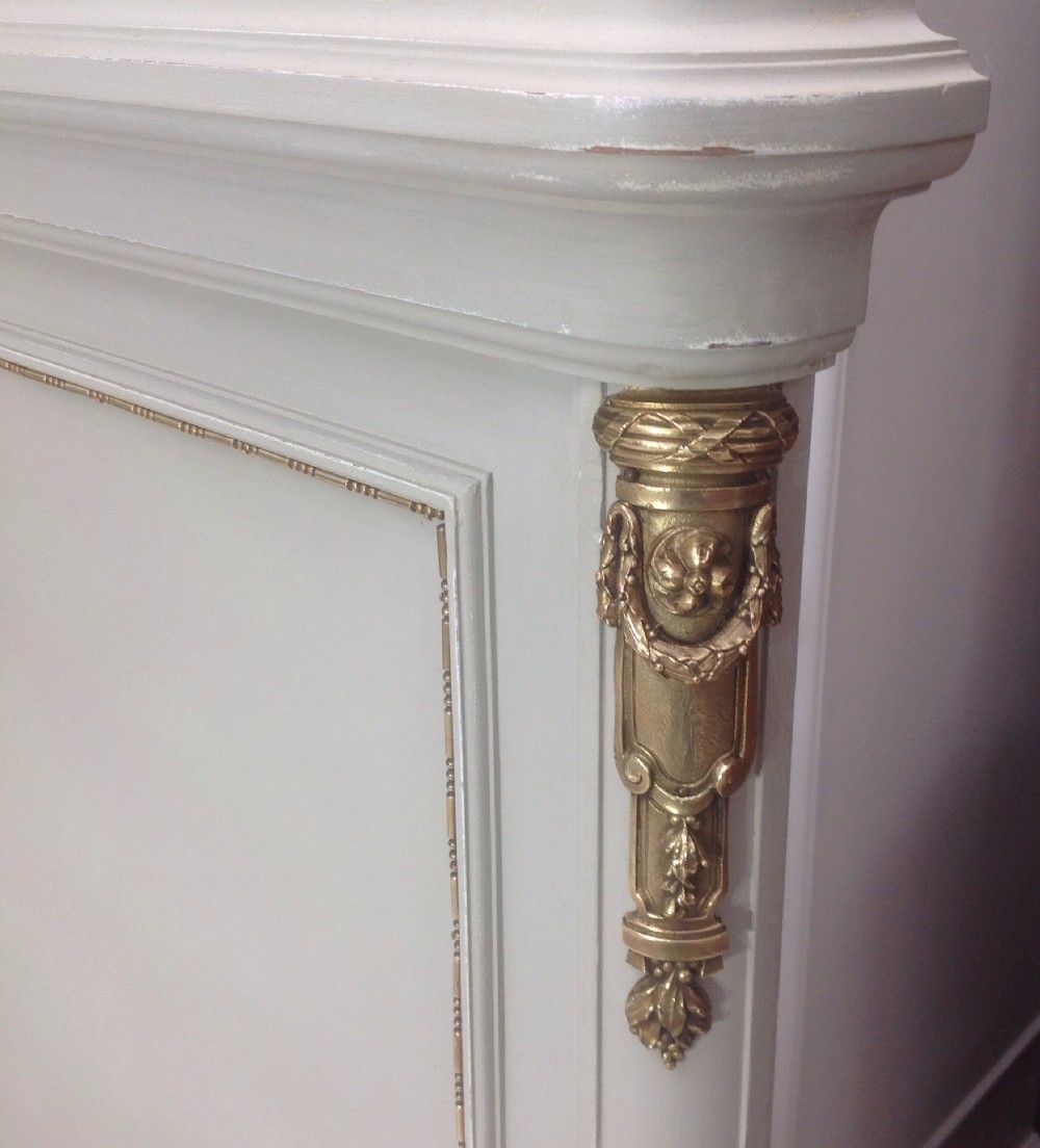An off-white painted French Neoclassical bed frame featuring a tall rectangular headboard, fluted turned legs, and intricate brass swag mounts on the footboard. View X