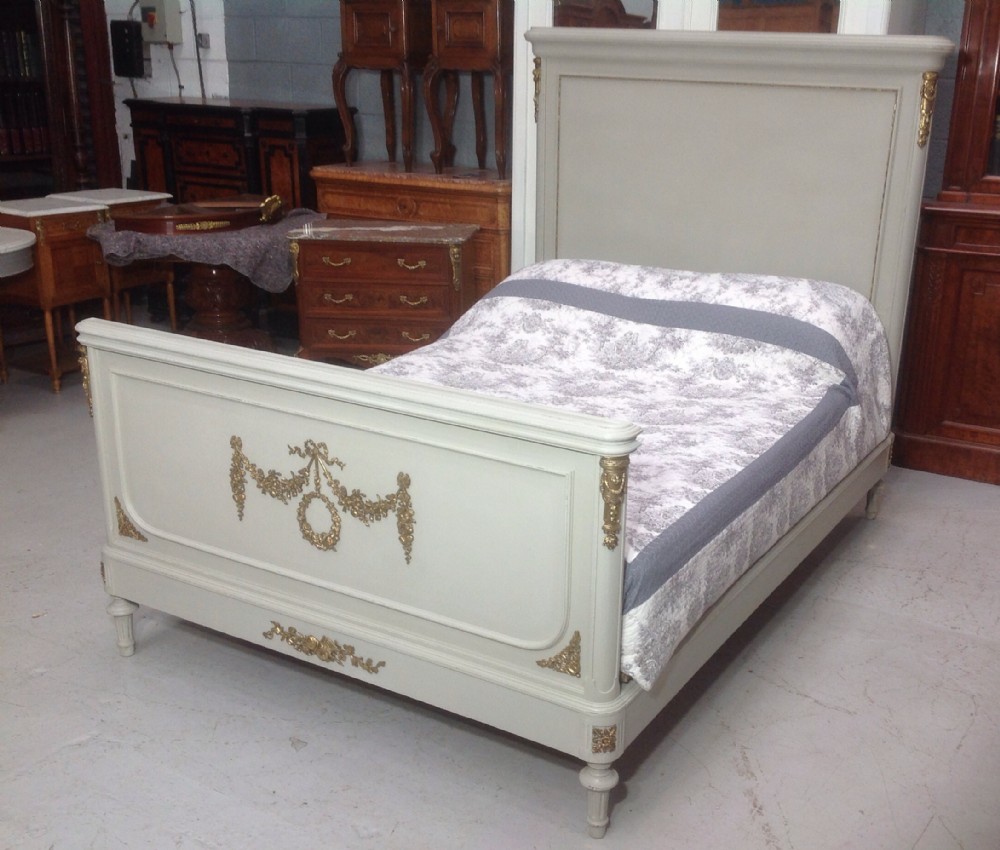An off-white painted French Neoclassical bed frame featuring a tall rectangular headboard, fluted turned legs, and intricate brass swag mounts on the footboard. View XIV