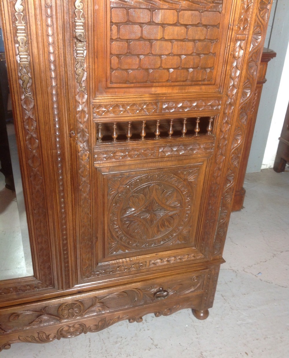 A tall carved walnut Breton armoire featuring one full-length mirrored door and one timber door with detailed relief carving and a spindle gallery. View III