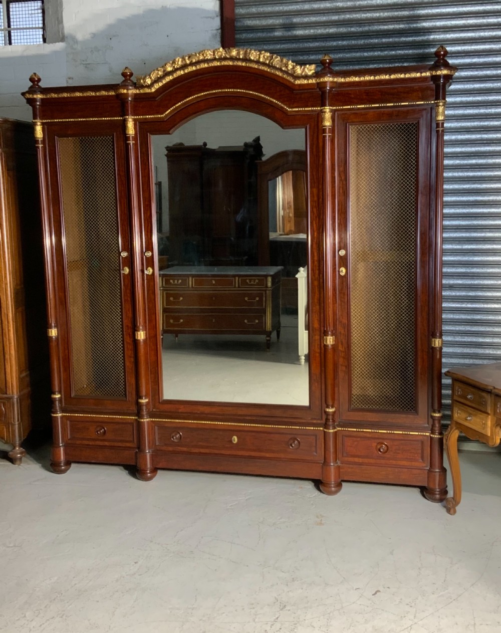 A large mahogany three-door armoire featuring a central arched mirrored door, wire-mesh side panels, and gilt bronze mounts.